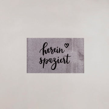 Gray washable doormat with "herein spaziert" slogan in black script, adds einzigartig Wohndeko and individueller Stil as a. - Waschbare Fußmatte Hereinspaziert